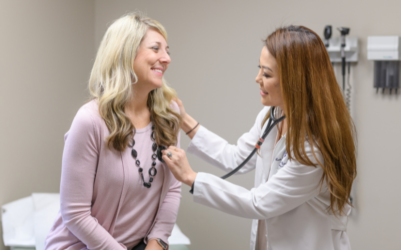 female provider with long brown hair listening to female patient's heart.